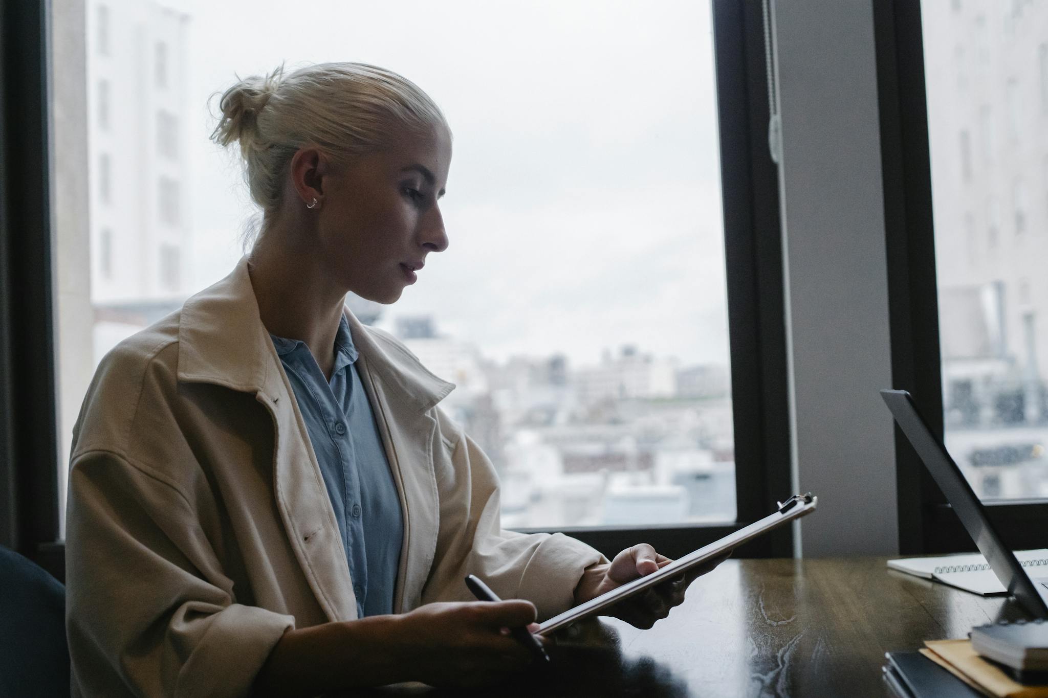 Side view of young concentrated female wearing white coat sitting at wooden desk in office and checking reports on clipboard